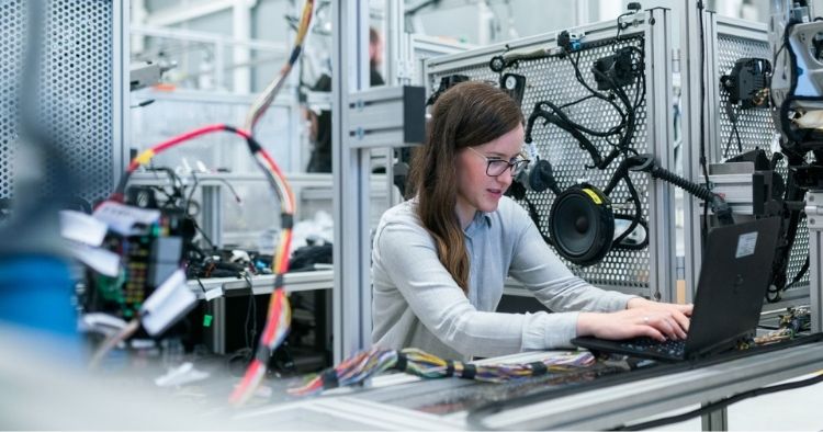 Woman working on a computer in a workshop surrounded by parts representing manufacturing companies